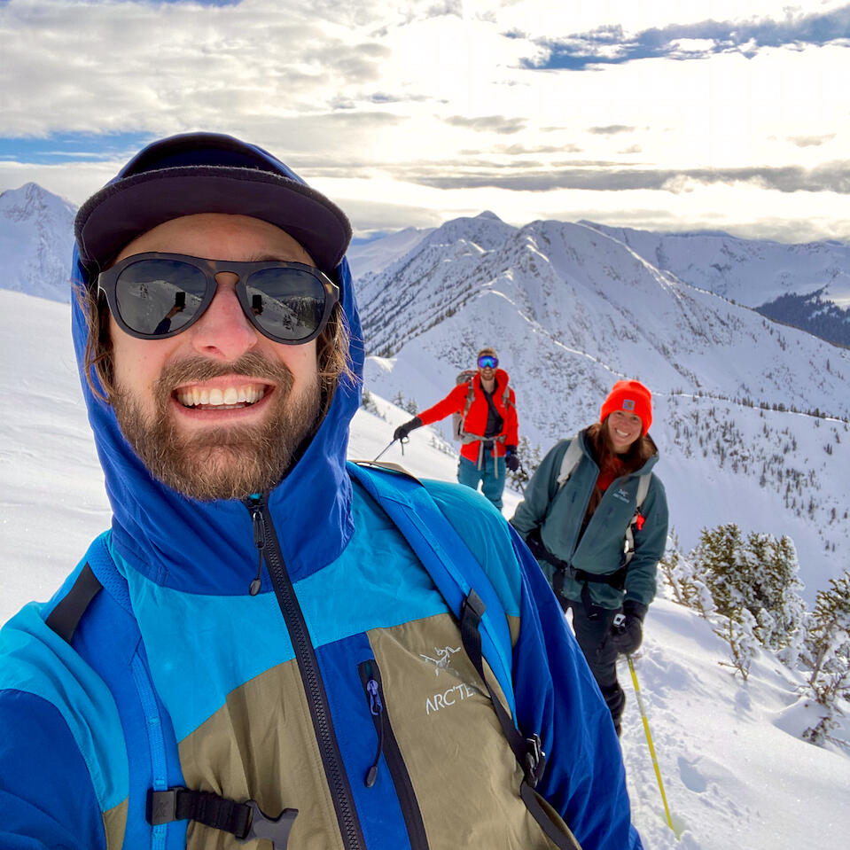 Devon de Balasi Brown on top of a mountain taking a selfie with friends in the background while ski touring and wearing a cap, sunglasses, blue jacket and backpack